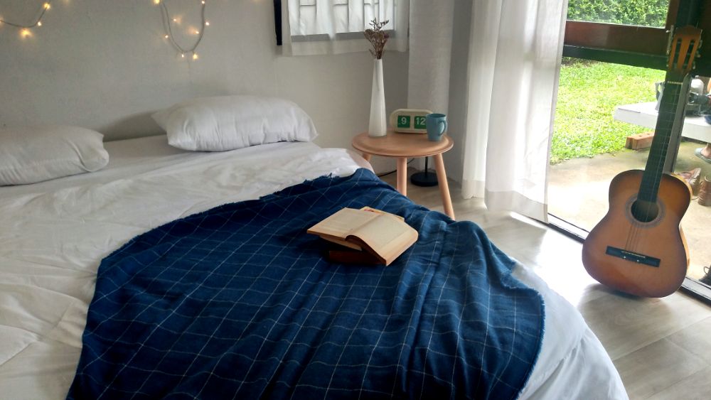 Foto de una habitación con una gran cama con sabanas blancas y una cobija azul, acompañado por una pequeña mesa de madera natural y una guitarra acústica.