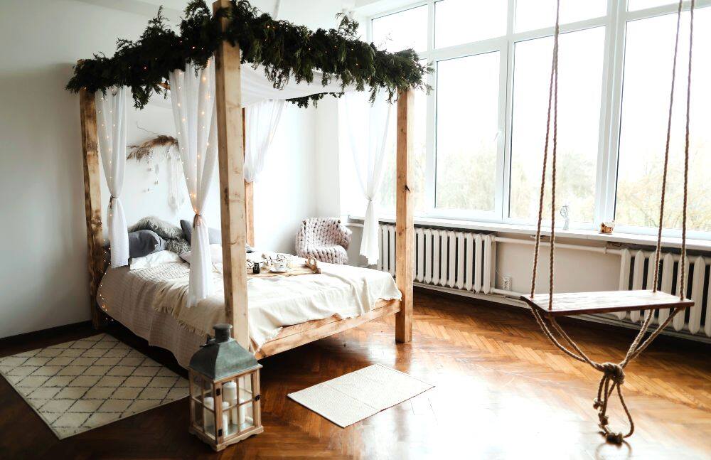 Un dormitorio con una cama rústica con dosel fabricada con madera natural y decorada con algunas plantas y cortinas blancas.