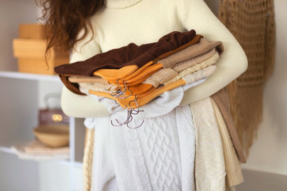La imagen muestra a una mujer sosteniendo un buen numero de sus suéteres y blusas, todos sostenidos por ganchos de madera.
