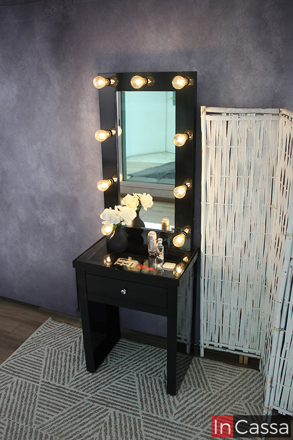 Mini tocador de color negro instalado en una pared gris sobre un tapete con patrón piramidal. El tocador incluye un espejo con iluminación LED integrada además de una cubierta de cristal que deja visible los maquillajes y utensilios que guarda el cajón.