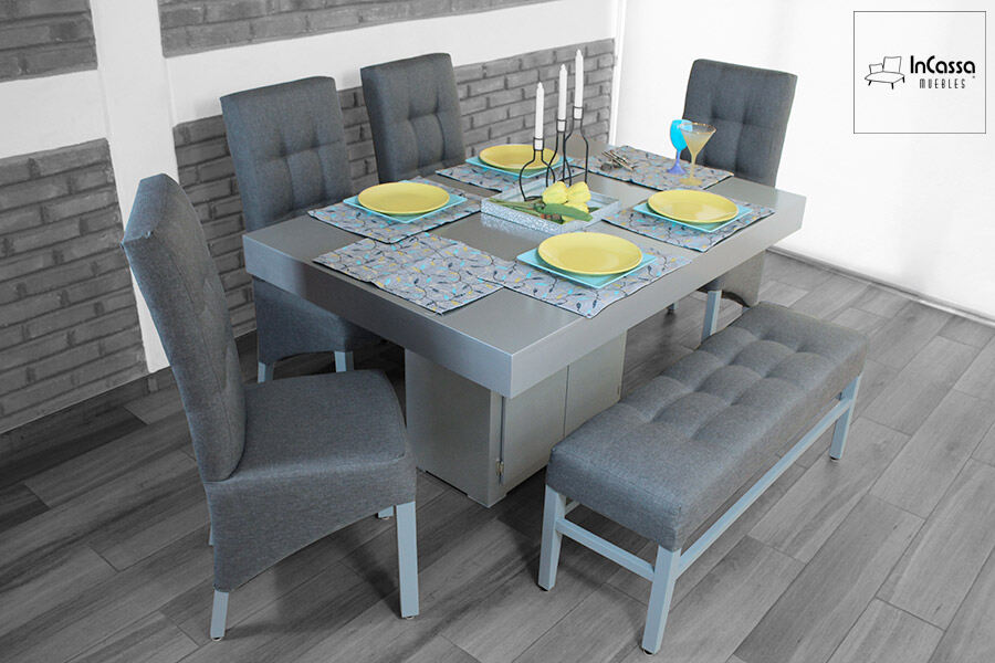 Comedor de 4 sillas y 1 banca para 2 personas. La mesa de color gris cuenta con compartimentos en el pedestal, mientras que las sillas y banca tienen un acabado capitoneado en su tapiz igualmente gris.