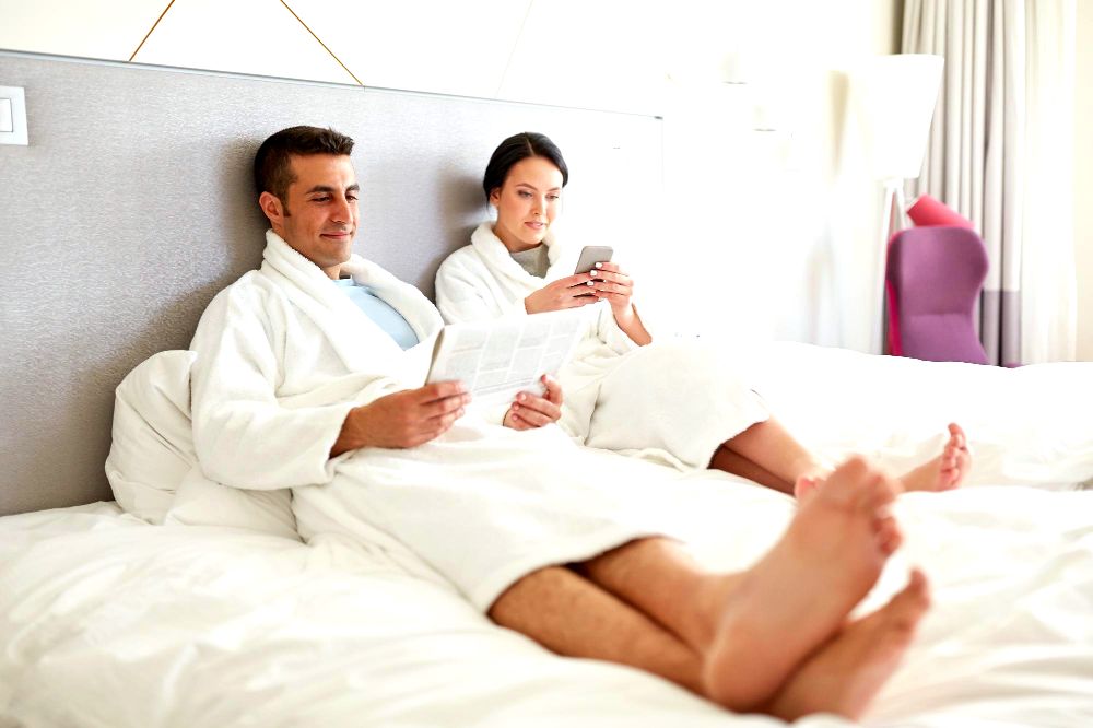 La imagen muestra a una pareja relajandose en su cama leyendo el periódico y revisando su teléfono móvil.