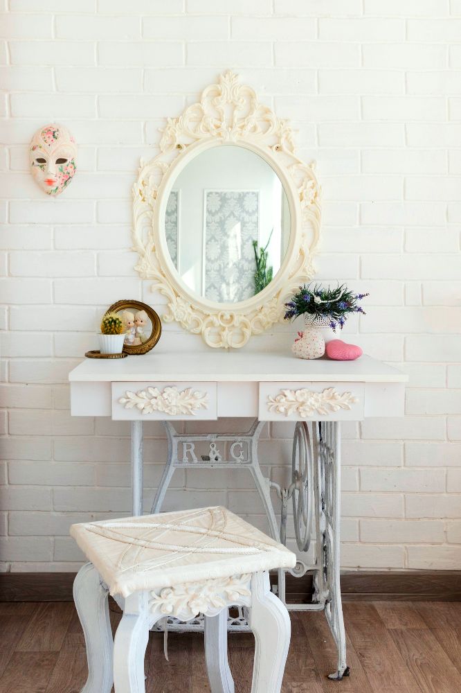 La foto muestra un hermoso tocador vintage de color blanco construido con madera y metal, diseñado con atractivos detalles tallados en sus cajones y en el marco de su espejo.
