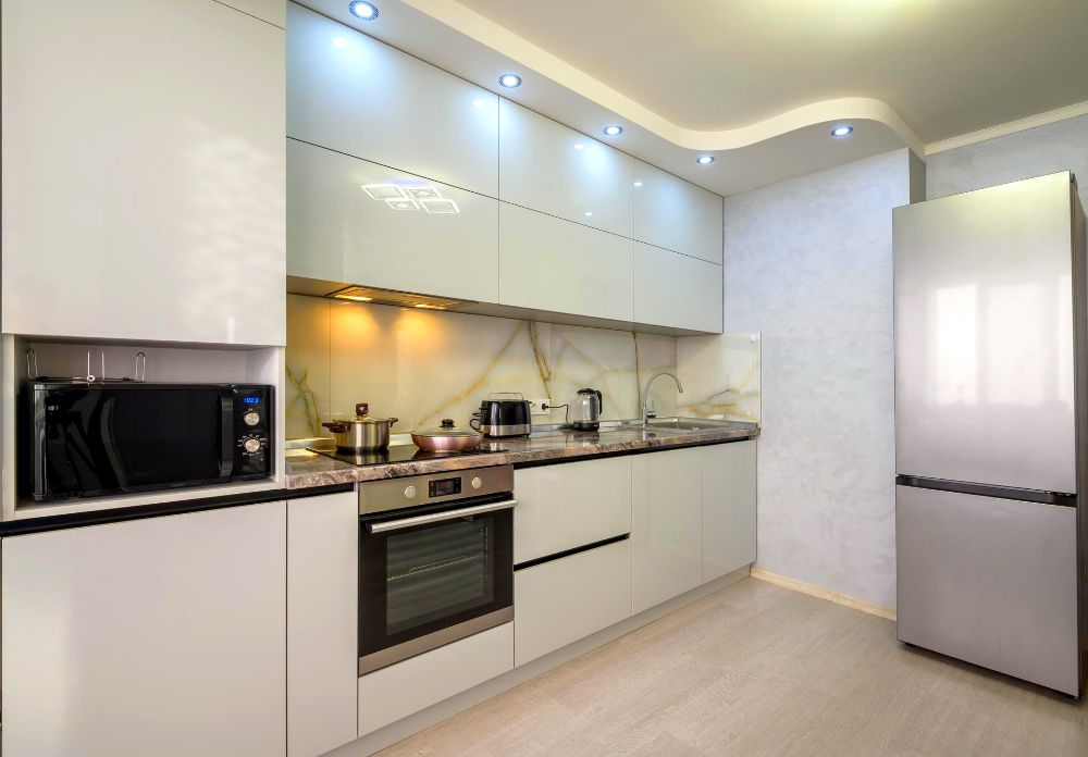 Foto completa de una cocina minimalista de color blanco, la cuál se encuentra acompañada de los electrodomésticos más esenciales, tales como microondas, refrigerador, parrilla entre otros.