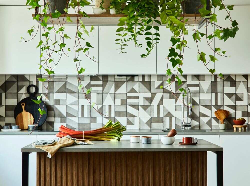 Cocina lineal de módulos blancos combinada con una isla de madera y cubierta de mármol, la toma frontal destaca los azulejos decorativos que decoran la cocina junto con un estante suspendido lleno de plantas decorativas.