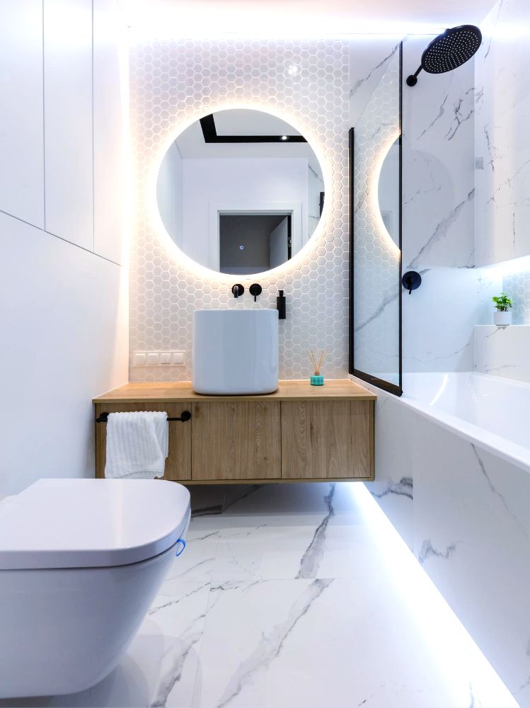 Una fotografía de un hermoso baño minimalista, en el que la totalidad de sus azulejos y piso es de tonos blancos dando más amplitud al espacio; el baño cuenta con taza y lavabo suspendidos, una amplia regadera y un espejo con iluminación en su marco.