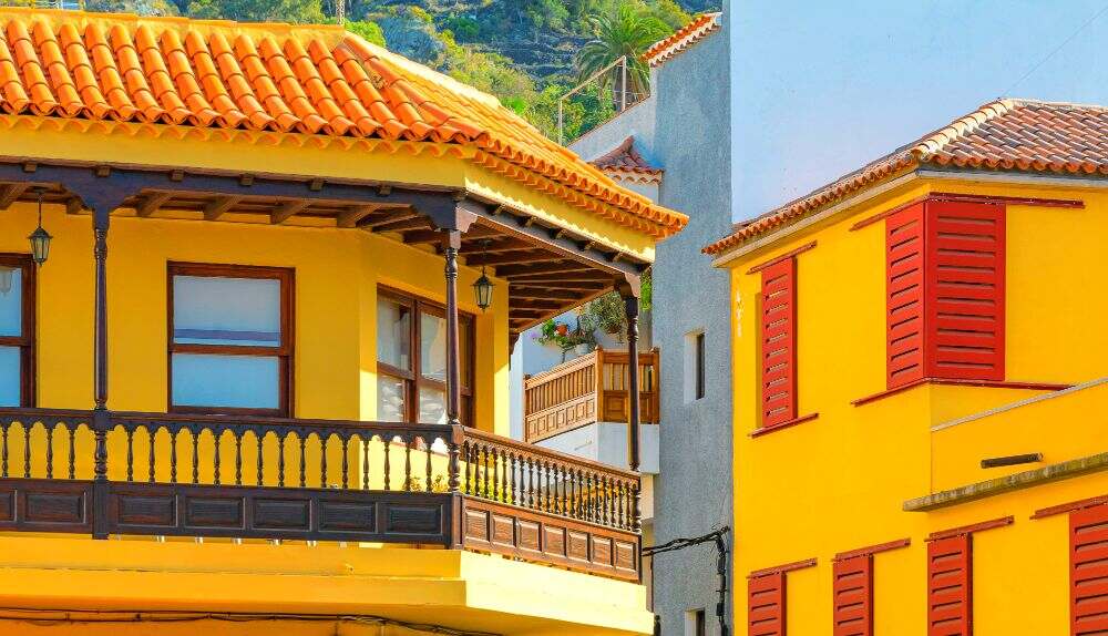 Una fotografía de los pisos superiores de dos casas cuyas fachadas son de color amarillo con algunos detalles en maple o naranja, lo que dota de alegría y vida al vecindario montañoso en el que se encuentran.