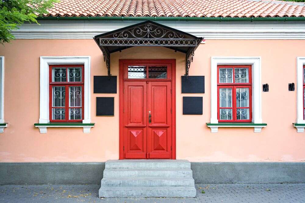 La foto presenta la fachada de una casa color salmón, en la que las ventanas y puerta son de color rojo con un marco de color blanco.