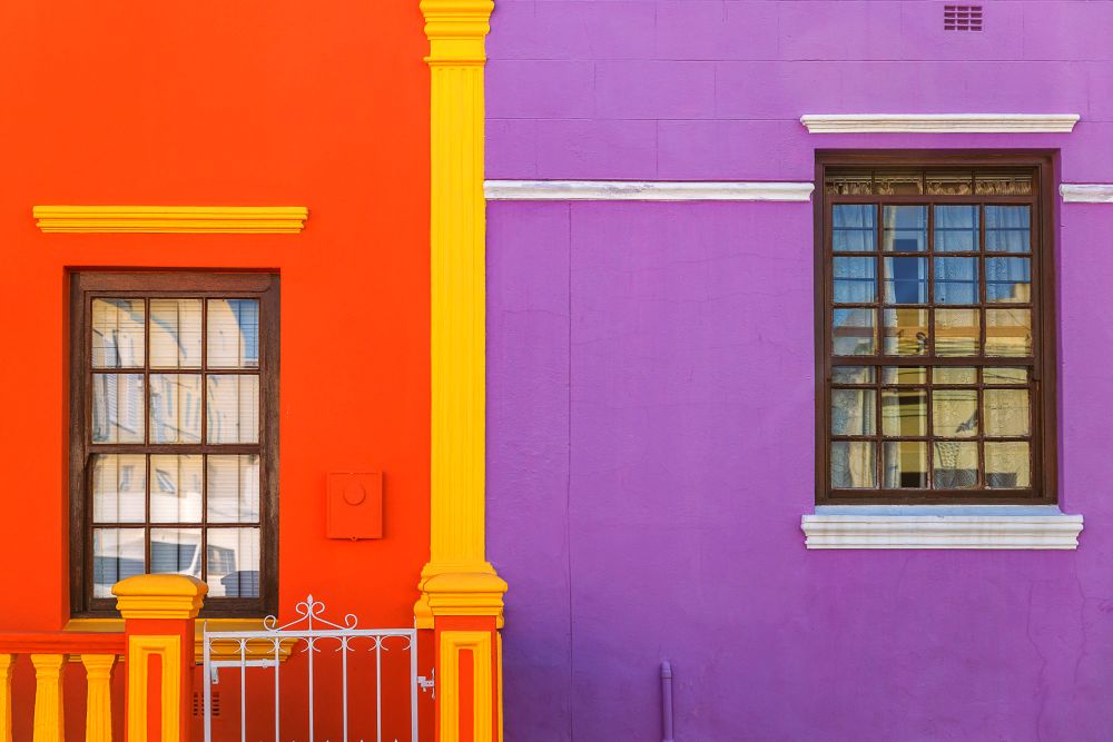 La imagen presenta la ventana de una casa de color naranja fuerte junto a la ventana de la casa vecina la cual es de color purpura.