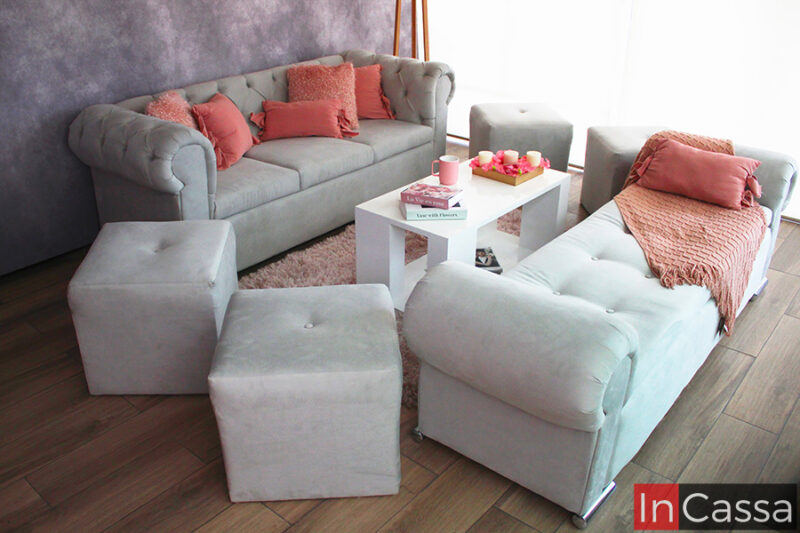 Sala tapizada en suede gris perla conformada por un sofá, un diván y 4 taburetes. La sala está decorada con algunos cojines y bordados rosas, y se encuentra presentada en una estancia con pared gris y piso de madera.