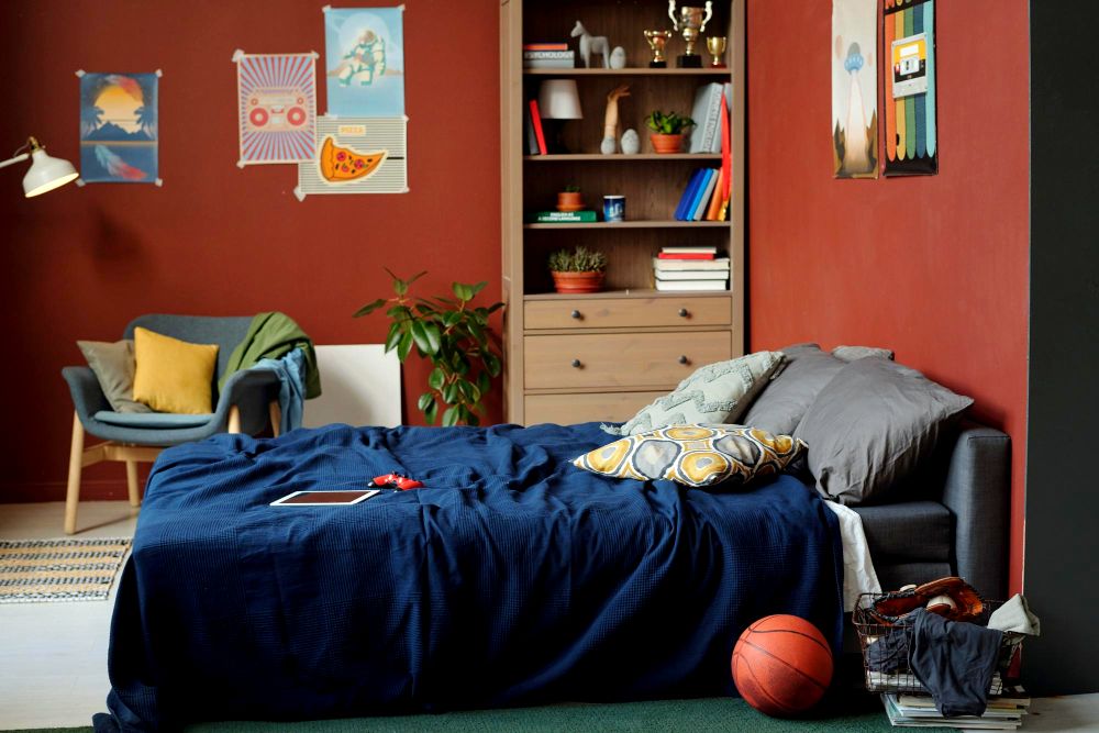 Una habitación con un estilizado estilo para muchachos, en la que vemos como cama a un futón de tapiz gris acompañada de un librero de madera natural y una silla minimalista. La habitación combina una decoración de tonos azulados con paredes rojas.