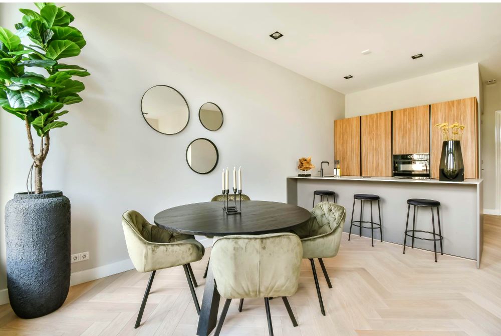 Un espacio abierto en el que se encuentra instalada una cocina minimalista con acabado natural y un comedor para 4 personas con mesa redonda negra y sillas capitoneadas aceitunadas. El lugar esta decorado con algunos espejos redondos de pared y una gran pl