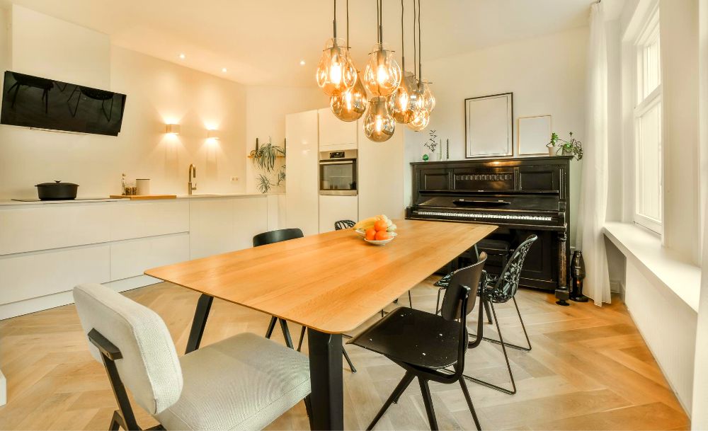 La foto presenta una cocina minimalista blanca con comedor de madera natural para 6 personas en un mismo espacio abierto, en el que resalta un piano clásico y unas lámparas flotantes de luz cálida que iluminan la estancia.