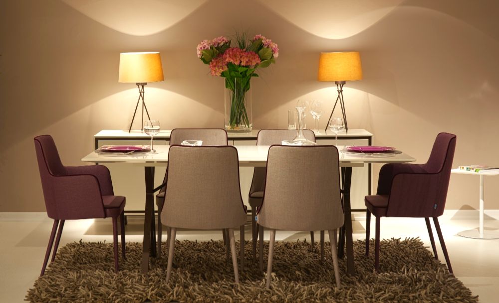 Fotografía de un comedor rectangular minimalista con 6 sillas modernas, la estancia además se encuentra decorada con dos lámparas minimalistas que proveen iluminación adicional y un gran florero de cristal con flores rosas. 