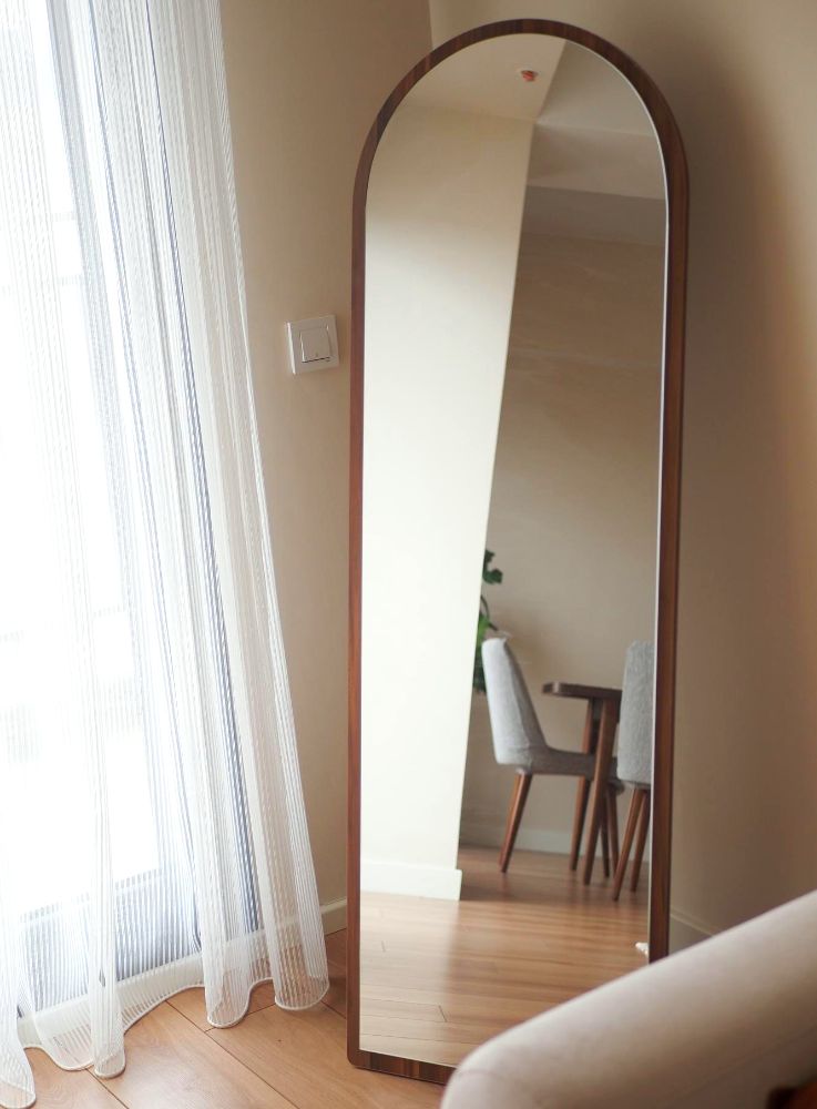 Foto de un espejo de cuerpo completo con marco arqueado de madera, instalado en una estancia junto a su ventana, lo que proporciona una sensación de amplitud y mayor iluminación al espacio.