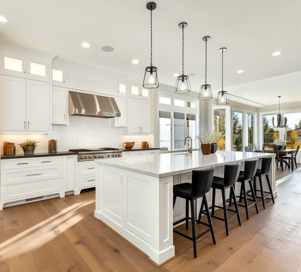 La foto presenta una amplia cocina con módulos, paredes y techo blancos, el ultimo con un gran sistema de iluminación en el que resaltan sus luminarias colgantes negras.