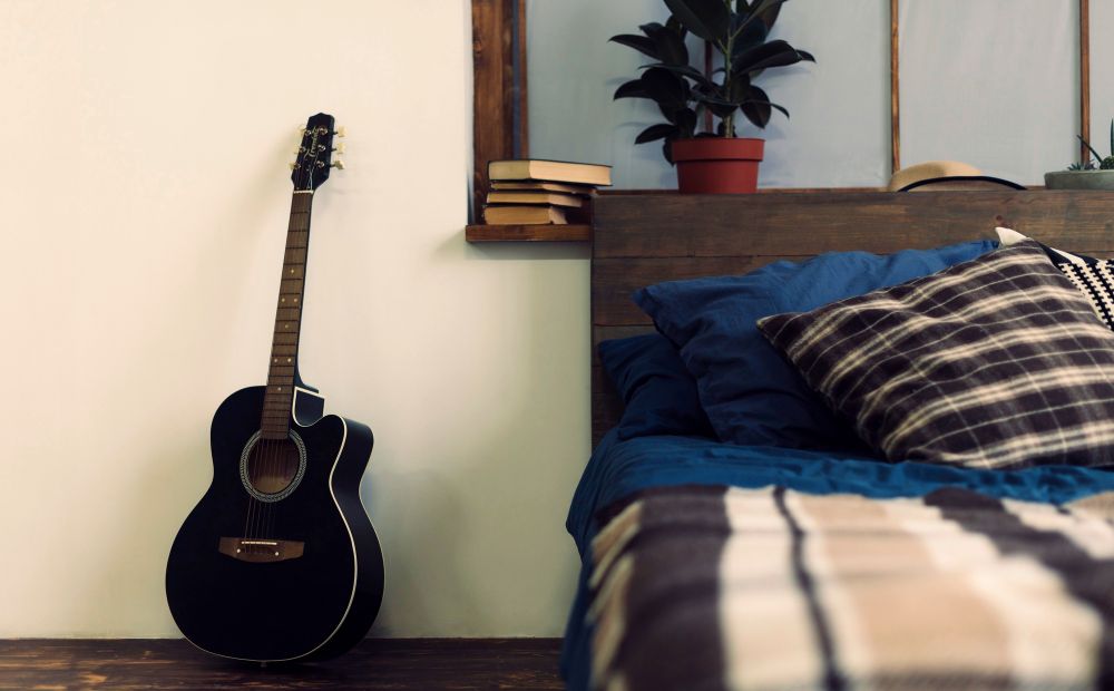 La imagen muestra una guitarra acústica apoyada en la pared junto a la cama de una habitación.