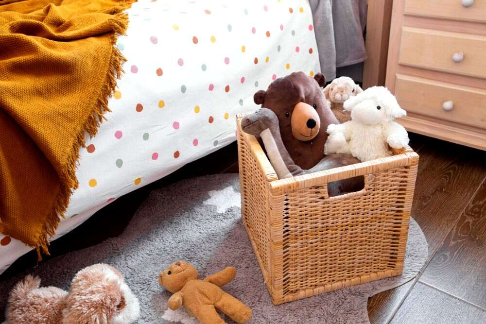 La foto muestra una caja de mimbre utilizada para guardar muñecos de peluche al lado de una cama.