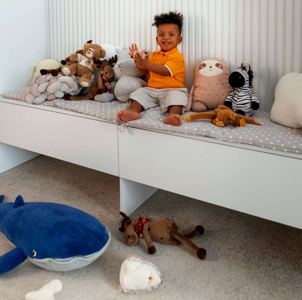 La foto muestra a un niño jugando con sus muñecos de peluche sentado sobre dos amplios baúles para juguetes, los cuales cuentan con almohadillas para servir también como asientos.