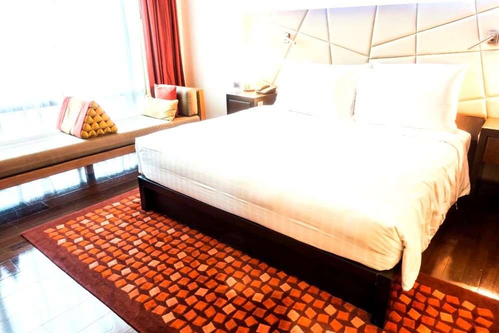 Moderna habitación con cama tamaño queen size, cojines coloridos y sofá junto a la ventana, sobre alfombra de patrones geométricos.