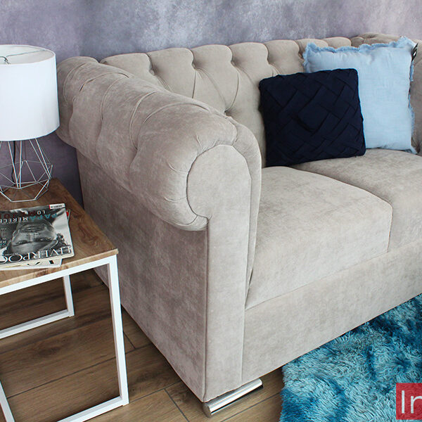 La foto presenta el love seat estilo capitoné en suede gris perla, acompañado de cojines azul oscuro y celeste. A su lado, una mesa auxiliar moderna sostiene una lámpara decorativa y una revista, complementando el ambiente sofisticado.