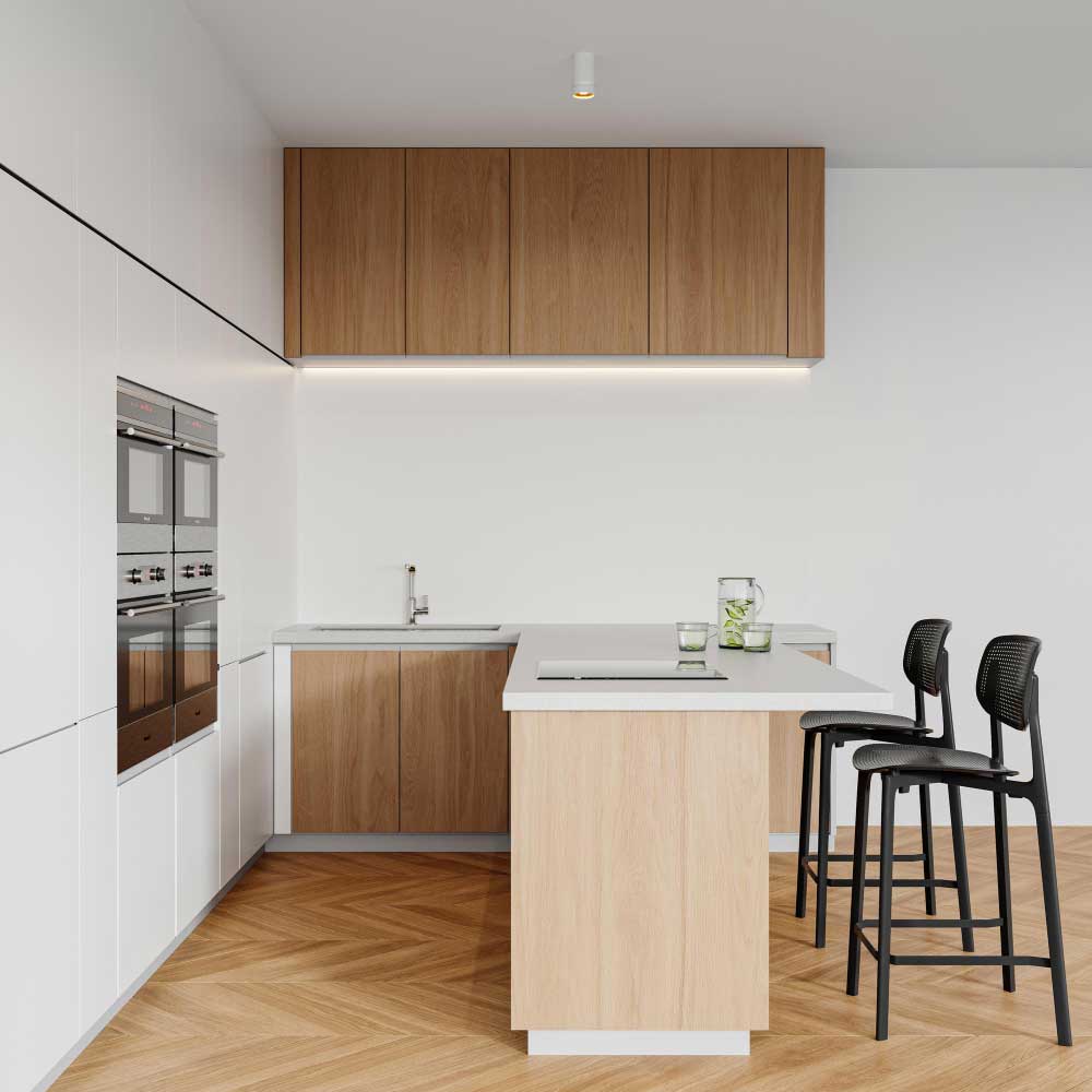 Una fotografía que presenta una cocina moderna en tonos cálidos con diseño minimalista. La isla central de madera clara con cubierta blanca integra una estufa empotrada y espacio para dos bancos altos negros. Al fondo, destacan los gabinetes superiores en madera y los inferiores en combinación madera-blanco, así como un elegante set de hornos empotrados en acero inoxidable. El piso en espiga y la luz tenue complementan el ambiente acogedor y contemporáneo.