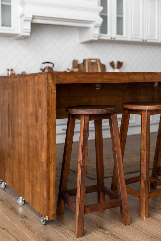 Una toma lateral cercana muestra una isla móvil fabricada en madera sólida con ruedas metálicas visibles en la parte inferior, lo que facilita su desplazamiento. Se acompaña de bancos altos del mismo tono cálido y rústico. Al fondo, se distingue una cocina de estilo clásico con gabinetes blancos, lo que crea un contraste acogedor y funcional entre lo tradicional y lo práctico.