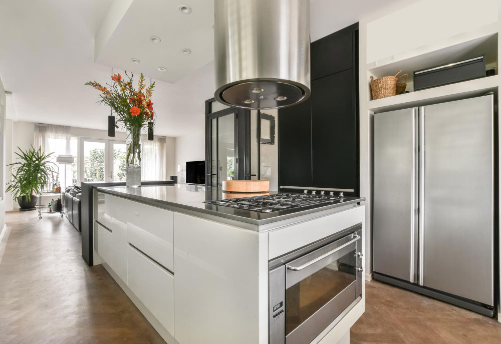La toma lateral destaca una isla moderna en acabado blanco brillante, equipada con horno, parrilla y una campana cilíndrica de acero inoxidable suspendida al centro. Sobre la cubierta, un florero con flores anaranjadas da un toque fresco al ambiente. Al fondo, se observa un refrigerador de doble puerta en acero y una sala luminosa que conecta con el jardín, creando una distribución abierta y funcional.