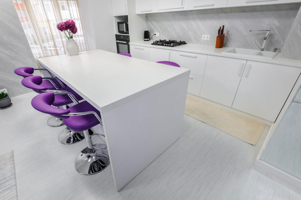 Una toma angular superior revela una moderna isla de cocina completamente blanca que contrasta con cinco banquitos altos en color morado vibrante. Esta combinación audaz añade personalidad al ambiente, mientras que el diseño limpio de líneas rectas mantiene un estilo minimalista. Al fondo, se observa una cocina lineal con cubierta blanca y salpicadero en acabado tipo mármol gris claro, equipada con parrilla empotrada, horno y tarja de doble tina. La luz natural refuerza el aire fresco y contemporáneo del espacio.