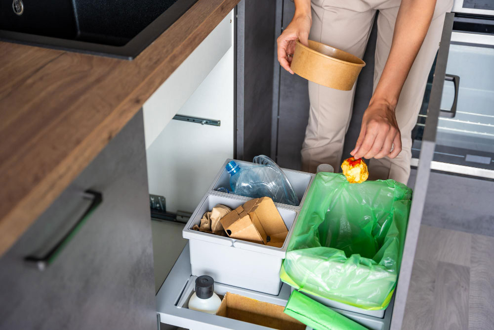Se presenta un práctico sistema de separación de residuos integrado dentro de una isla de cocina, captado en vista frontal mientras una persona deposita desechos. El módulo cuenta con compartimentos extraíbles que facilitan el reciclaje de basura orgánica, plásticos y cartón. La cubierta de madera oscura y los frentes en acabado gris mate aportan un estilo moderno y funcional, ideal para cocinas que priorizan la organización y sostenibilidad.