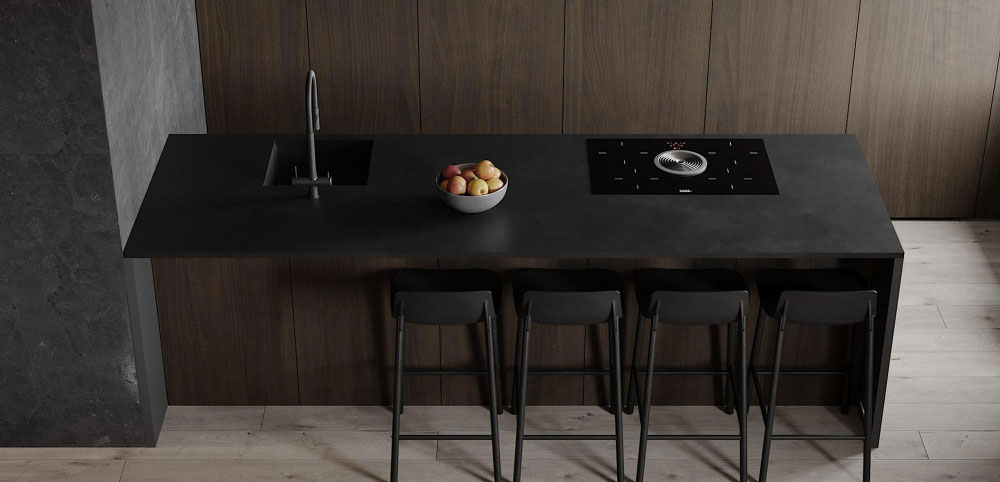 Una toma desde arriba muestra una cocina sofisticada y elegante, donde predomina la isla negra con cubierta mate, equipada con fregadero de acero negro y parrilla vitrocerámica con extractor central. Al frente, se alinean cinco bancos altos en tono oscuro que complementan la estética monocromática. El fondo en madera oscura y el contraste con el piso claro aportan un balance perfecto entre modernidad y calidez.
