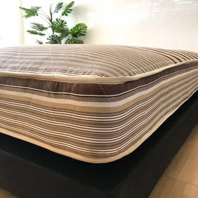 Vemos el colchón hotelero, con un estilo de rayas en el acolchonado, presentado sobre una base tapizada negra en una habitación con pared blanca y piso de madera.