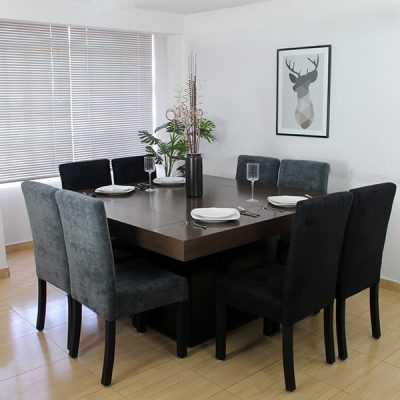 Comedor para 8 personas con 4 sillas tapizadas en suede gris y 4 sillas de suede negro, mientras que la mesa del comedor es de MDF chileno en acabado tabaco. EL comedor es presentado en una estancia con paredes blancas.