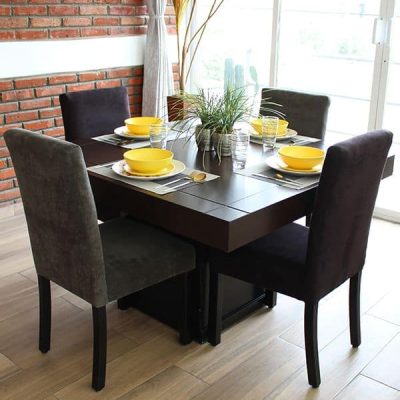 Imagen del comedor en una práctica presentación de 4 sillas, cuya mesa es cuadrada con una cubierta en acabado tabaco y un pedestal de color chocolate, mientras que sus sillas están tapizadas en suede en tonos grises y chocolate. El conjunto se exhibe en una habitación con pared de ladrillo y piso de madera.