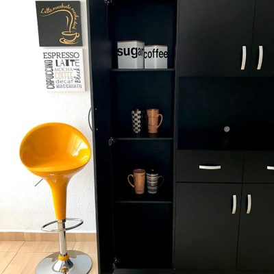 La foto presenta la Alacena para Microondas MÁLAGA en acabado chocolate mate con la puerta lateral izquierda abierta, mostrando tres repisas internas organizadas con frascos de azúcar y café, así como tazas decorativas. El contraste lo aporta un banco alto amarillo brillante junto a la pieza.