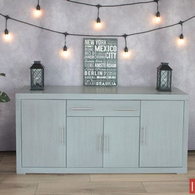 La imagen presenta el Bufetero Vintage Modelo MISSISSIPPI en un elegante tono gris cenizo claro, capturado de frente para resaltar su composición simétrica y líneas rectas. Se aprecian sus tres puertas amplias y dos cajones superiores con jaladeras metálicas alargadas, así como su acabado liso y moderno. Al fondo, un ambiente decorativo con luces cálidas y elementos minimalistas complementa perfectamente el estilo del mueble.