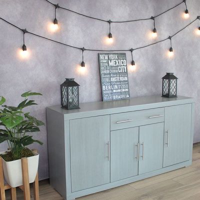 La imagen muestra una toma lateral del Bufetero Vintage Modelo MISSISSIPPI en color gris cenizo claro, destacando su acabado homogéneo y textura suave. El ángulo resalta la profundidad del mueble, sus puertas amplias con jaladeras metálicas y los cajones superiores perfectamente alineados. La escena decorada con luces cálidas, una planta y faroles negros crea un ambiente acogedor que realza su estilo moderno y elegante.