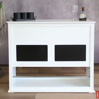 Moderna Cantina blanca con parches decorativos negros y barra posa pies metálica incorporada. El mueble es presentado junto a un fondo de tono gris y piso de madera.