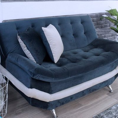 La fotografía presenta el love seat del modelo Murcia, tapizado en suede azul marino con detalles en tono hueso. Su estructura robusta y diseño capitonado destacan sobre un fondo neutro con muros de ladrillo gris y plantas naturales, acentuando su estilo moderno y su elegancia atemporal.