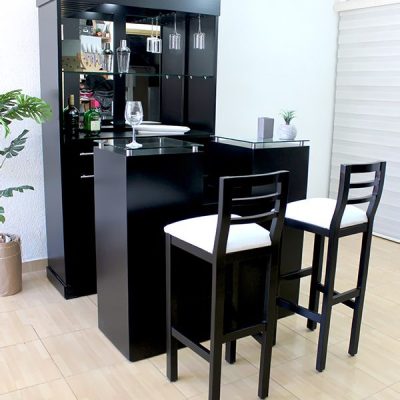 La imagen muestra una elegante cantina en acabado chocolate mate, vista en ángulo frontal. El mueble cuenta con una barra principal, repisas de vidrio, soporte para copas colgantes y espejo trasero que aporta profundidad. Se acompaña de dos bancos altos con estructura de madera en negro y asientos tapizados en blanco, ideales para complementar el diseño.